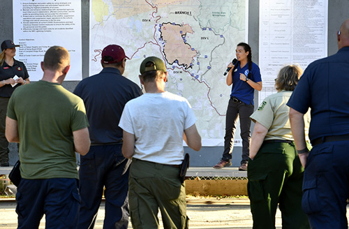 A group of firefighters and forest personnel attend a wildfire incident briefing in front of a large topographic map, where a woman with a microphone provides an operational update.
